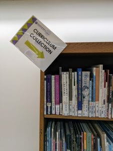 Sign set at an angle, reads "Curriculum Collection" with an arrow pointing down to the right and a bookshelf.
