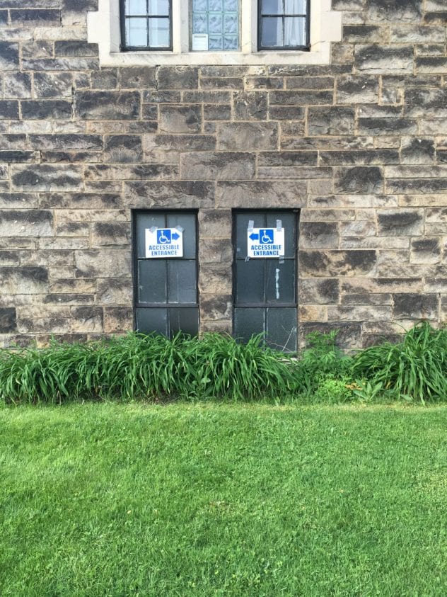 Image of a stone church wall with two small windows showing an accessibility sign with arrows pointing to the left, and the other sign pointing to the right with conflicting directions.